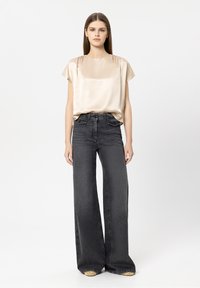 Beiges Satintop mit kurzen Ärmeln, kombiniert mit hochtaillierten, dunkelgrauen Jeans mit weitem Bein. Auffälliges goldenes Detail an den Schuhen.