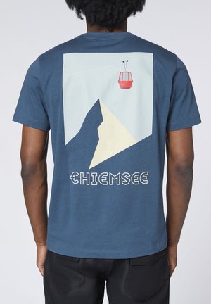 Person wearing a blue t-shirt with a mountain and red cable car design and the word "Chiemsee" on the back.
