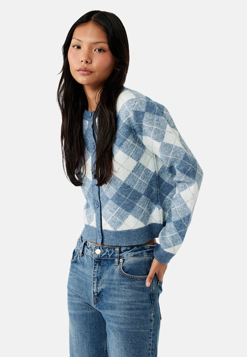 Jonge vrouw met lang donker haar draagt een blauw-witte argyle cardigan en blauwe spijkerbroek, poserend met één hand in de zak.
