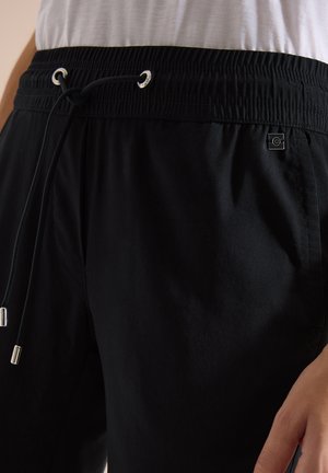 Černé kalhoty s elastickým pasem a šňůrkami s kovovými konci a malou čtvercovou nášivkou s logem u kapsy, nošené s bílým topem.