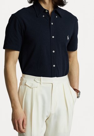 Mann trägt ein eng anliegendes schwarzes Kurzarm-Hemd mit weißem Logo, cremefarbene hochgeschnittene plissierte Hose und eine Armbanduhr.