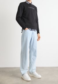 Felpa nera con logo "Calvin Klein", indossata sopra una camicia a righe con colletto. Jeans dritti azzurri e scarpe da ginnastica bianche completano l'outfit.