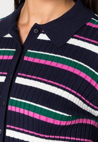 Marinblå ribbad pikétröja med horisontella ränder i rosa, vitt och grönt. Inkluderar krage och knapppdetaljer.