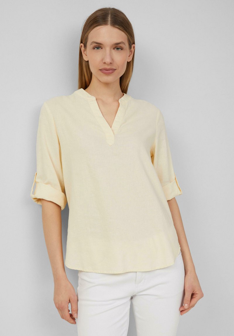 Blouse col boutonnière jaune clair avec des manches amples retroussées. Tissu doux et texturé avec un ourlet arrondi. Design simple et minimal.