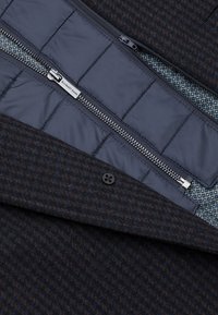 Płaszcz z wełny w mieszance z ciemno-brązowym wzorem w kratkę; posiada pikowaną podszewkę, zamek błyskawiczny oraz niebieską geometryczną podszewkę wewnętrzną.