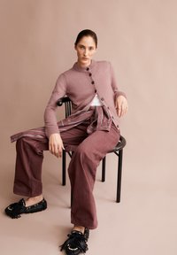 Frau, seitlich auf einem schwarzen Stuhl sitzend, trägt eine mauvefarbene Strickjacke, passende weite Hosen, ein am Bund gebundenes gestreiftes Hemd und schwarze Quasten-Liedenschaftsschuhen.