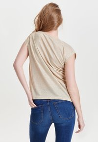 Kvinde med lysbrunt hår iført en glitrende beige kortærmet top og blå jeans, vist fra ryggen med den ene hånd i lommen.