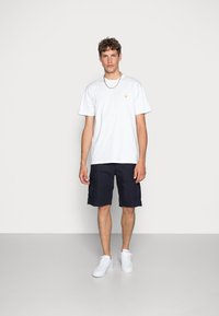 White cotton t-shirt with a small yellow logo, paired with dark navy cargo shorts and white sneakers. Minimalistic design and casual fit.