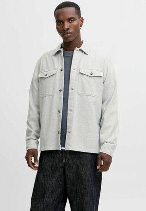 Camisa de franela gris claro con dos bolsillos en el pecho, botones negros y un ajuste holgado, superpuesta a un suéter azul con textura. Jeans oscuros usados por debajo.