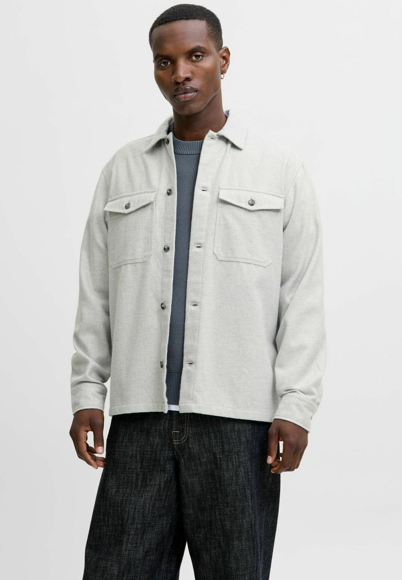 Camisa de franela gris claro con dos bolsillos en el pecho, botones negros y un ajuste holgado, superpuesta a un suéter azul con textura. Jeans oscuros usados por debajo.