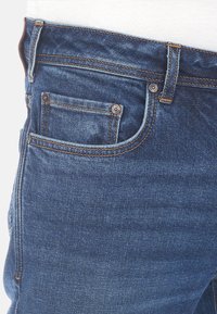 Jeans de mezclilla azul oscuro con un diseño clásico de cinco bolsillos, que presentan remaches de cobre, costuras naranjas y una textura suave.