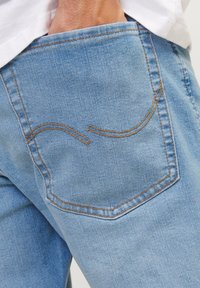 Lys blå denimjeans med visning av en baklomme med buet søm i oransje. Stoffet er mykt med en lett tekstur.