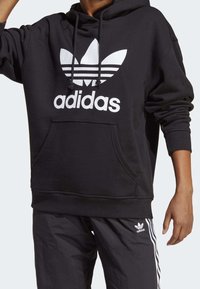 Svart huvtröja i mjukt tyg, med en stor vit Adidas-logga och tre horisontella ränder, samt en framficka i kattunge-stil.