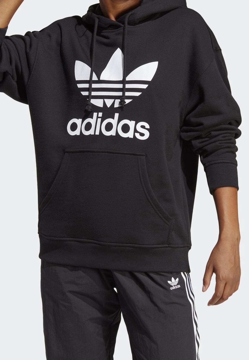 Svart huvtröja i mjukt tyg, med en stor vit Adidas-logga och tre horisontella ränder, samt en framficka i kattunge-stil.