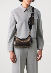 Sac en bandoulière en cuir serpent métallisé brun avec des accessoires dorés, de forme courbée et avec une bandoulière amovible. Porté sur une tenue à carreaux noir et blanc.