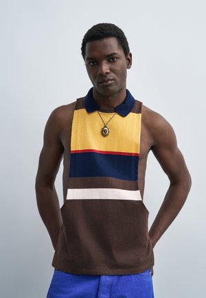 Bărbat care poartă un tricou polo fără mâneci cu dungi orizontale galbene, bleumarin, maro, albe și roșii și pantaloni albaștri, stând pe un fundal simplu.