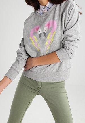 Personne portant un sweat-shirt gris avec des têtes de flamants roses et des éclairs jaunes, associé à un pantalon vert clair.