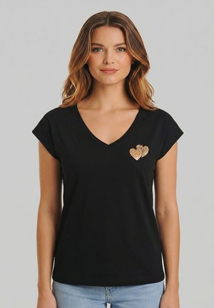 Vrouw met golvend bruin haar die een zwart T-shirt met V-hals draagt, met twee gestructureerde hartvormige ontwerpen op de linkerborst, gecombineerd met lichtblauwe spijkerbroek.