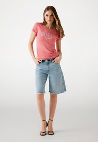 Rožinė trumpa rankovė t-shirt su „GUESS EST.1981“ logotipu, derinama su šviesiai mėlynomis džinsinėmis Bermuda šortais su suplėšytu apvadu ir juodu diržu. Juodos aukštakulnės šlepetės.