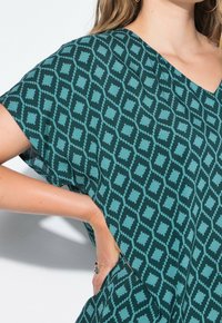 T-shirt ample de couleur sarcelle avec un motif géométrique aux nuances sombres et claires. Le tissu a une texture douce et légère.