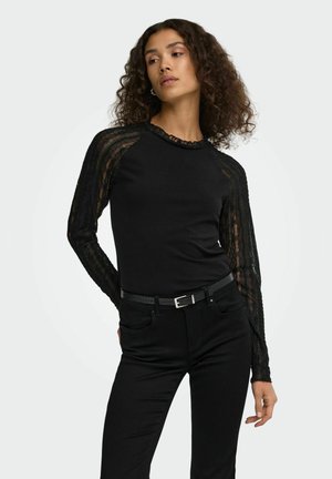 Femme aux cheveux bouclés portant un haut noir à manches longues en dentelle et un pantalon noir avec une ceinture, posant avec une main dans le dos.