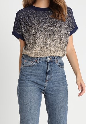 Vrouw draagt een kortemouwen marineblauw en beige gebreide top met vlekken, ingestopt in een high-waisted blauwe spijkerbroek.