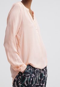 Blusa rosa chiaro con placket a bottoni, maniche lunghe e vestibilità morbida, abbinata a pantaloni scuri con motivo a foglie verdi.