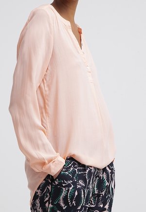 Blusa rosa chiaro con placket a bottoni, maniche lunghe e vestibilità morbida, abbinata a pantaloni scuri con motivo a foglie verdi.