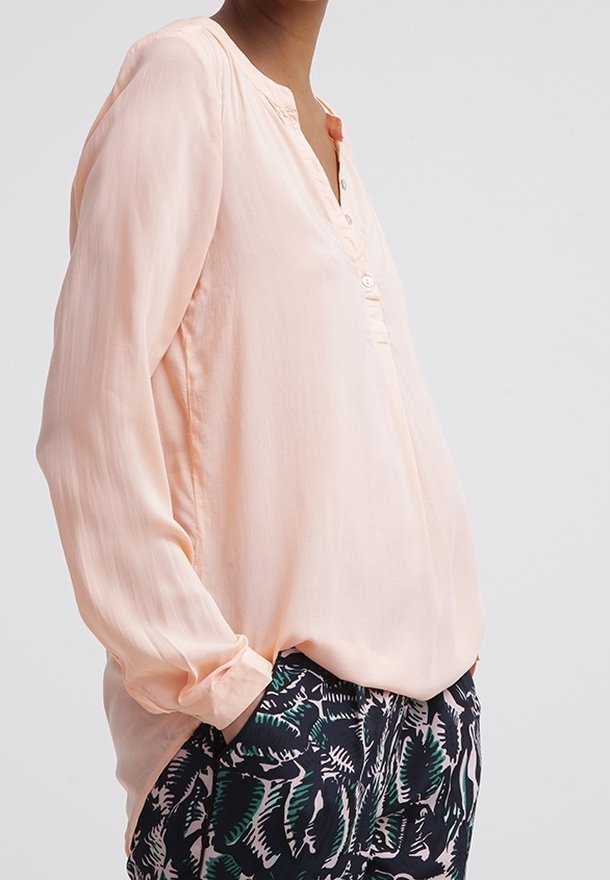 Blusa rosa chiaro con placket a bottoni, maniche lunghe e vestibilità morbida, abbinata a pantaloni scuri con motivo a foglie verdi.