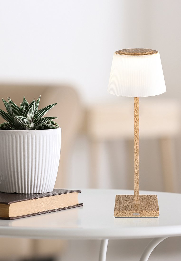 Lampada da tavolo in legno con paralume bianco plissè, base cilindrica e supporto quadrato in legno, posizionata accanto a una pianta in vaso su un tavolo bianco.