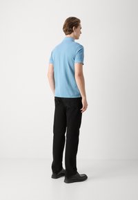 Tricou polo de culoare albastru deschis, cu mâneci scurte, asortat cu pantaloni negri drepti și pantofi negri. Vedere din spate.