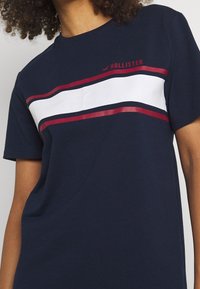 Granatowa bawełniana koszulka z krótkim rękawem, z białymi i czerwonymi poziomymi pasami, z logo "Hollister" w kolorze czerwonym nad pasami.