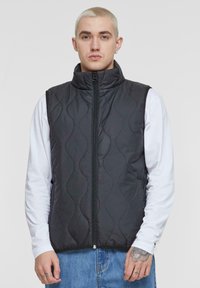 Schwarze gesteppte Weste mit hohem Kragen und Frontreißverschluss, ausgestattet mit einer glatten Textur und einem geschwungenen Nähmuster, getragen über einem weißen Langarmshirt.
