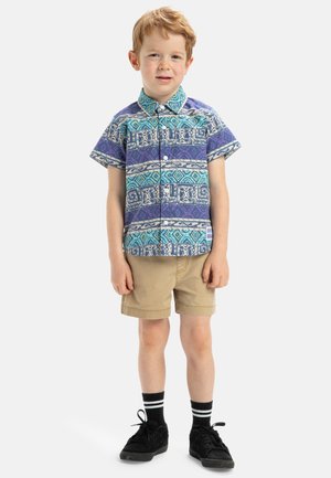 Kurzärmliges Hemd mit geometrischem Muster in Blau, Lila und Weiß, kombiniert mit beigen Shorts und schwarzen Turnschuhen mit gestreiften Socken.