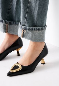 Zapato de tacón negro de cuero con puntera afilada, acento en forma de triángulo dorado y tacón metálico corto y cuadrado. Lleva puesto con jeans de mezclilla arremangados.