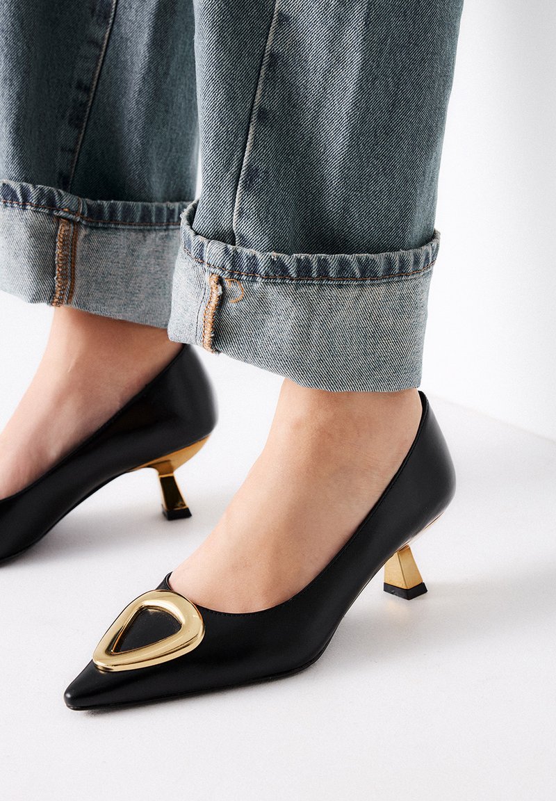 Zapato de tacón negro de cuero con puntera afilada, acento en forma de triángulo dorado y tacón metálico corto y cuadrado. Lleva puesto con jeans de mezclilla arremangados.