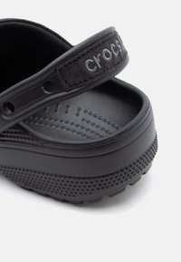 Close-up of black Crocs clog showing textured footbed, heel strap with embroidered logo, and patterned sole edge.