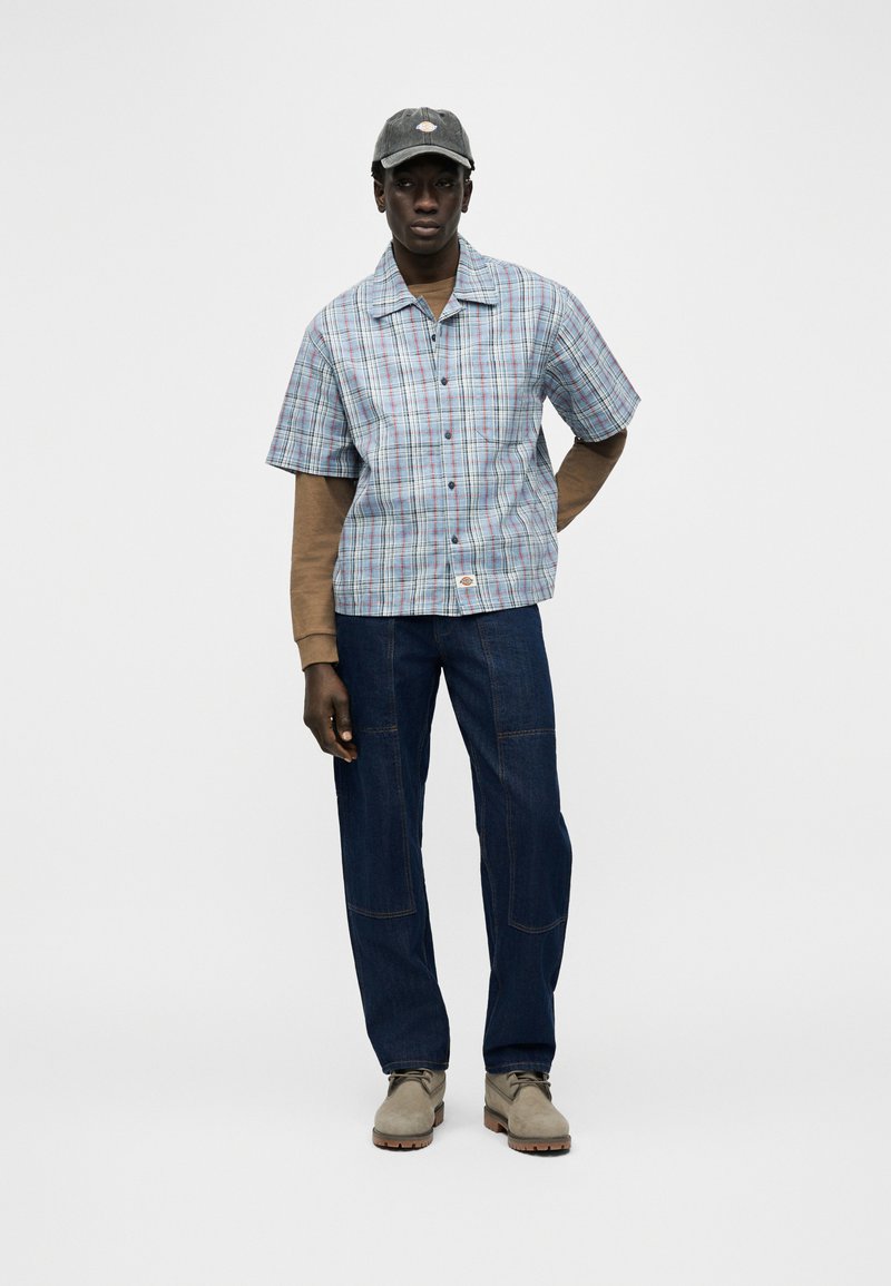 Mann mit grauer Kappe, hellblau kariertem Kurzarmhemd über braunen Langarmshirts, dunkelblauer Jeans und beigen Schuhen, steht mit einer Hand hinter dem Rücken.