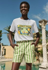 Balta marškinėliai su spalvinga "Amalfi Coast Lemons" grafika, derinami su žaliais ir baltais dryžuotais šortais su šoninėmis sagtimis. Lauko aplinka.