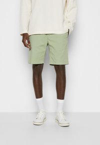 Pantaloni corti di cotone verde chiaro con fronte piatto e tasche laterali, abbinati a calze bianche e scarpe da ginnastica bianche casual.