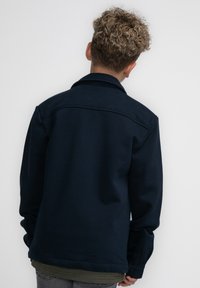 Donkerblauwe jacket met een kraag, knoopmanchetten en rechte snit. Gemaakt van een textuurstof, gedragen over een groene blouse, weergegeven vanuit de rug.