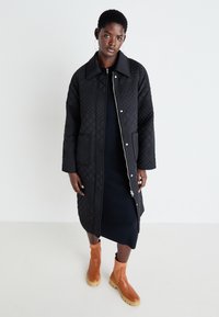 Manteau long noir matelassé avec un col prononcé, doté d'une fermeture éclair et de pressions. Porté par-dessus une robe sombre et assorti à des bottines brunes.