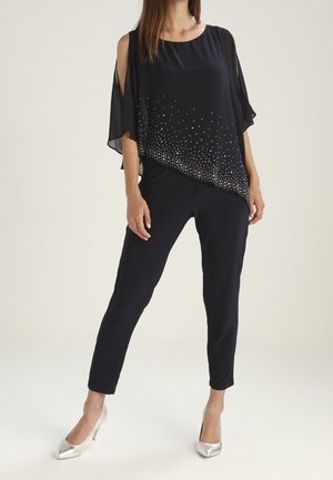 Haut noir transparent à épaules dénudées avec des ornements argentés, associé à un pantalon noir ajusté et des talons argentés brillants.