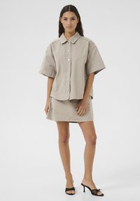 Beige oversized shirt with large collar and button closure, paired with a matching mini skirt featuring decorative studs. Black heeled sandals.