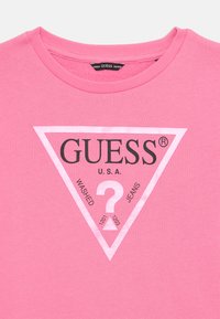 Camiseta de algodón rosa con un gráfico triangular que incluye el texto "GUESS®", "U.S.A." y "WASHED JEANS" en negro, con un signo de interrogación central.