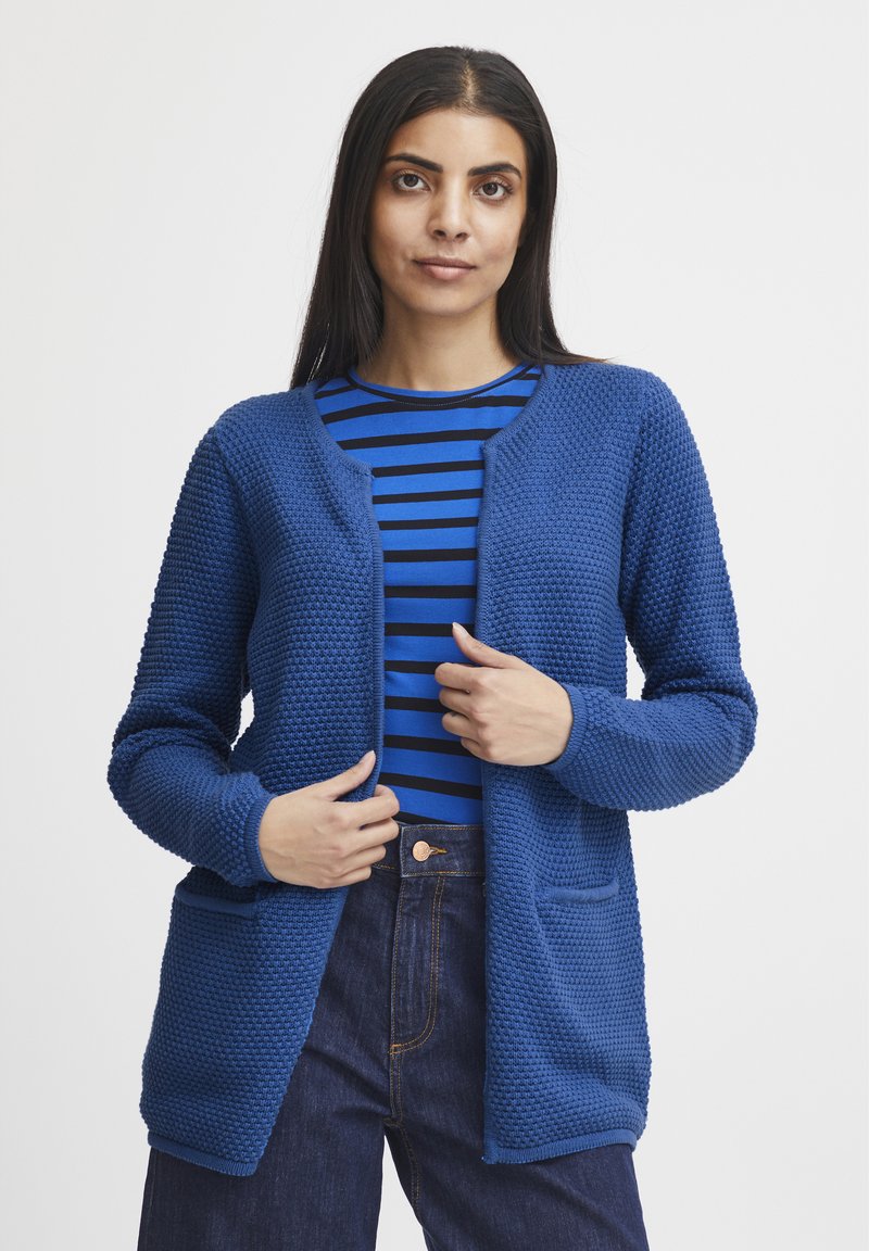 Blauer gestrickter Cardigan mit strukturiertem Muster, offener Vorderseite und zwei vorderen Taschen. Getragen über einem blau-schwarz gestreiften Oberteil.