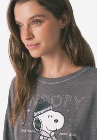 Grijze T-shirt met lange mouwen, met daarop een zwart-wit Snoopy-afbeelding met een hoed. Zacht materiaal, ronde hals en decoratieve stiksels.