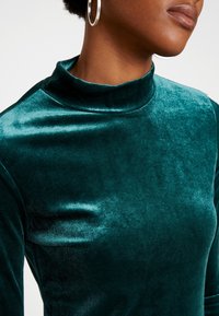 Suéter de terciopelo verde azulado con una textura suave y un diseño ajustado, que presenta un cuello alto y mangas largas. Pendientes de aro plateados visibles.