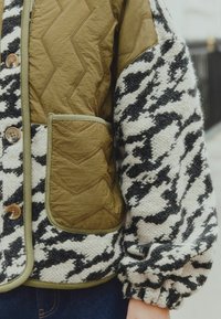 Kudumistehnikaga oliivroheline kangas, millel on chevron-õmblus ja eesmine tasku, koos musta ja valge mustriga tekstureeritud varrukatega. Ümmargused nupud on nähtavad.