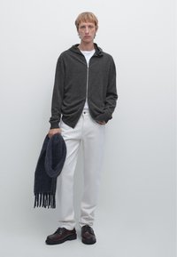 Mörkgrå zip-up hoodie, vit t-shirt under, vita byxor, håller en mörkblå scarf. Bruna skor med strukturerad sula.